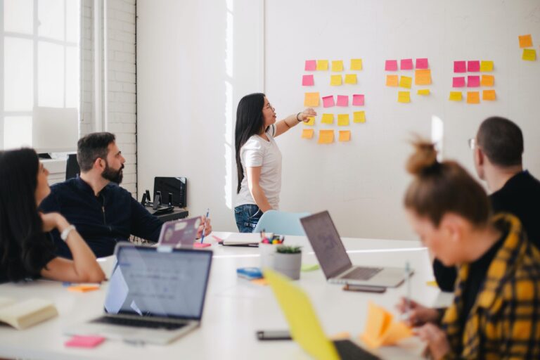 A woman leads a meeting, pointing at colourful sticky notes on a whiteboard, while four colleagues sit at a table with laptops and papers.