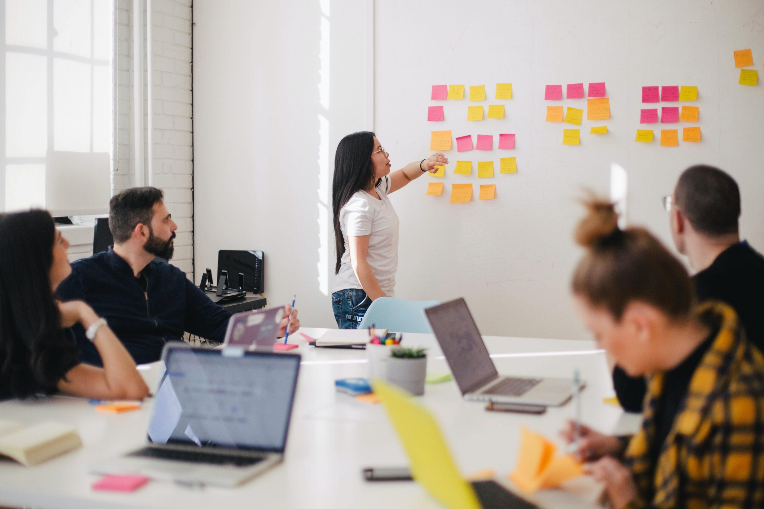A woman leads a meeting, pointing at colourful sticky notes on a whiteboard, while four colleagues sit at a table with laptops and papers.
