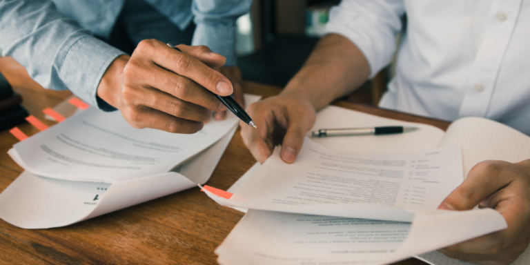 Two people sit at a wooden desk, reviewing and discussing documents with tabs. One person points at a page with a pen whilst the other holds the papers open. A pen rests on the table beside them.