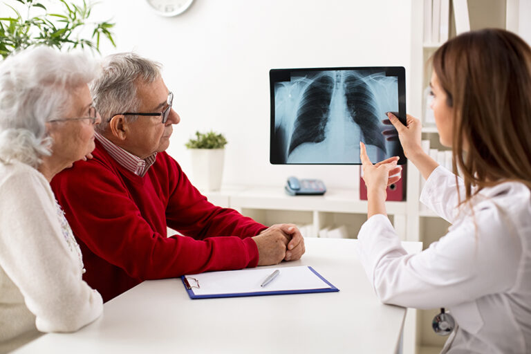 A doctor shows an elderly couple a chest X-ray on a tablet during a medical consultation at a desk, with a clipboard and medical equipment visible.
