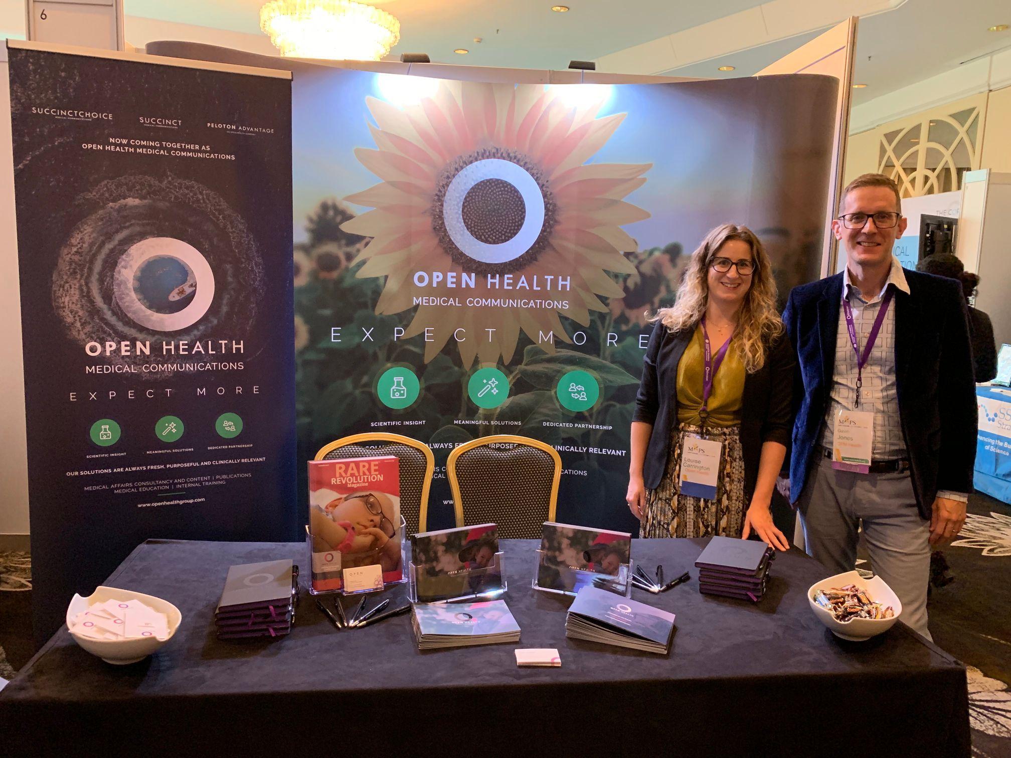 Two people stand behind a table at an OPEN Health Medical Communications stand, featuring banners, brochures, and a sunflower graphic at a conference or event.