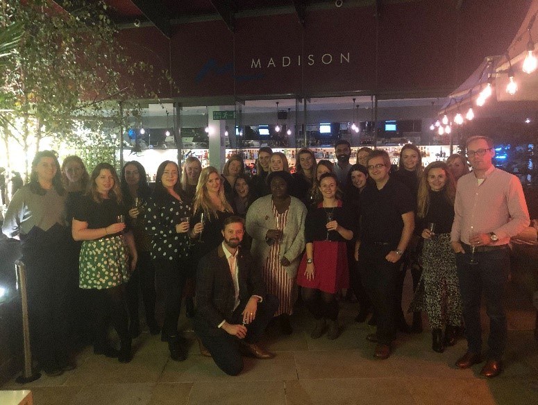 A group of about 24 people pose together indoors at night, some holding drinks, with fairy lights above and a Madison sign in the background.