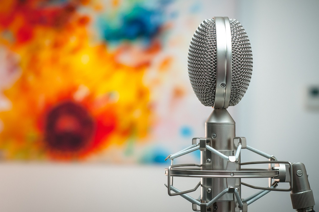 A silver vintage-style microphone stands in front of a blurred, colourful abstract painting with orange, yellow, blue, and red hues.