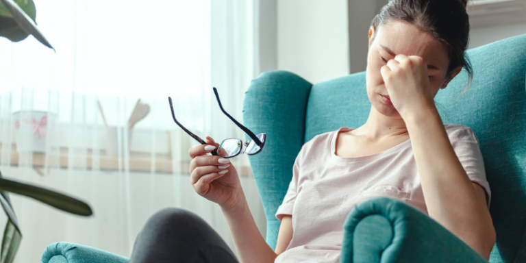 A woman sits in a teal armchair, holding her spectacles in one hand and pinching the bridge of her nose, appearing tired or stressed.