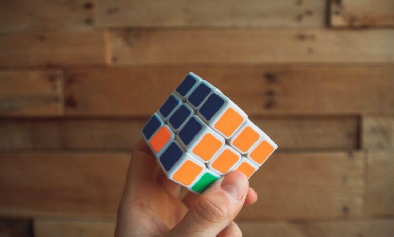 A hand holds a partially solved Rubik's Cube with orange, blue, green, and white stickers, against a wooden background.