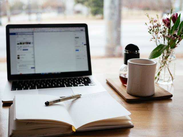An open laptop sits on a wooden desk beside a notebook with a pen, a mug on a tray, a small vase with flowers, and a window showing an outdoor scene in the background.