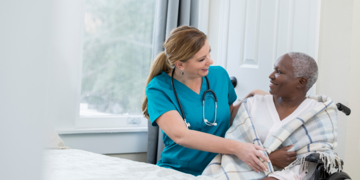 A nurse in blue scrubs smiles whilst covering an older woman with a blanket. The woman sits in a wheelchair near a bed, and both appear happy in a bright, cosy room.