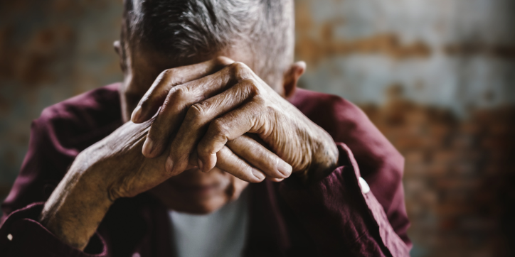 An elderly person with grey hair rests their head on folded hands, partly covering their face, wearing a maroon shirt. The background is blurred, creating a sombre and reflective atmosphere.