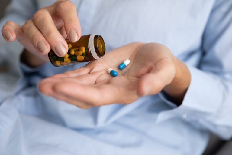 A person pours yellow and green capsules from a brown bottle into their hand, which already holds two blue and white capsules.