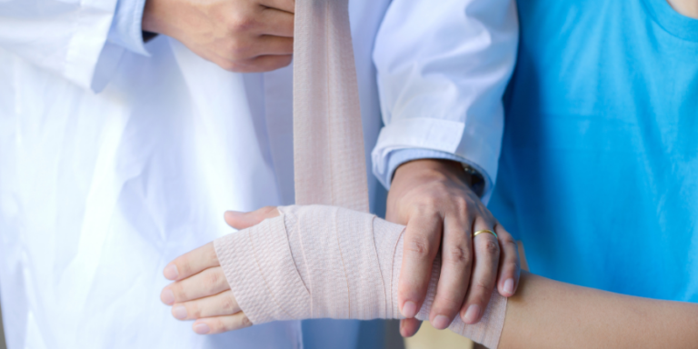 A doctor in a white coat wraps a beige bandage around a patient's injured wrist while gently supporting the patient's hand.