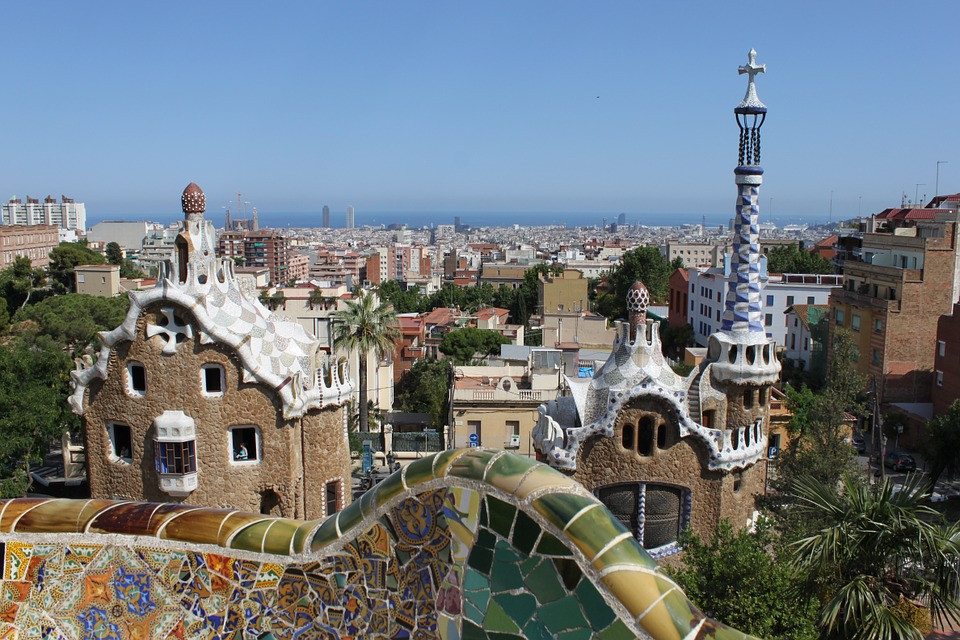 View of colourful mosaic tiles, whimsical buildings, and cityscape in Park Güell, Barcelona, with Mediterranean Sea in the background under a clear blue sky.