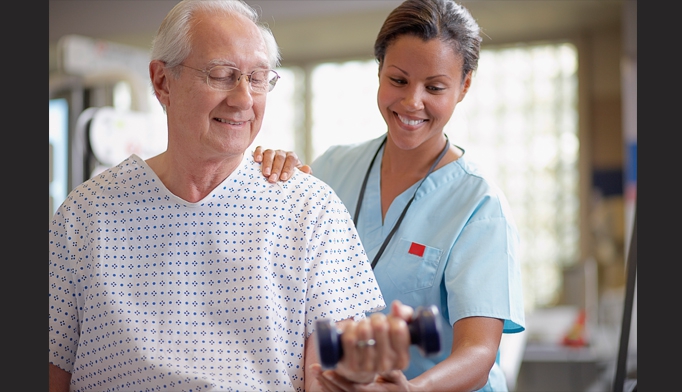 Smiling nurse helps an elderly man in a hospital gown lift a small dumbbell during a physiotherapy session. They are indoors, and both appear happy and engaged in the activity.