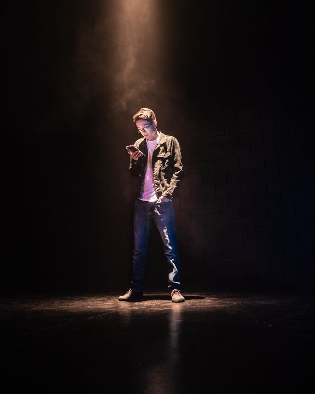 A person stands alone on a dark stage under a spotlight, looking at a mobile in their hand. They wear jeans, trainers, and a jacket.