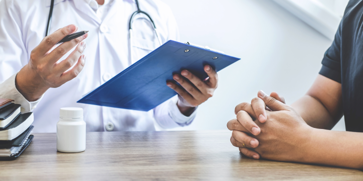 A doctor holding a clipboard discusses something with a patient, who sits across the table with hands clasped. A pill bottle and books are on the table between them.