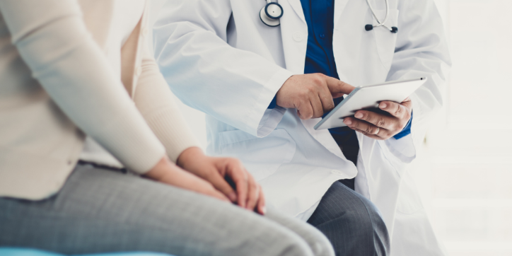 A doctor in a white coat holds a tablet and talks with a patient sitting nearby. The focus is on their hands and torso, not their faces.