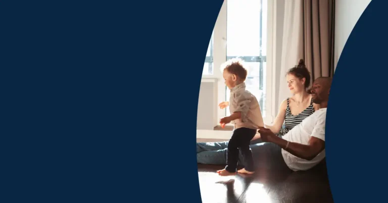 A family with a young child sits and plays together on the floor by a sunny window. The image is partially bordered by curved navy blue shapes on the left and right sides.