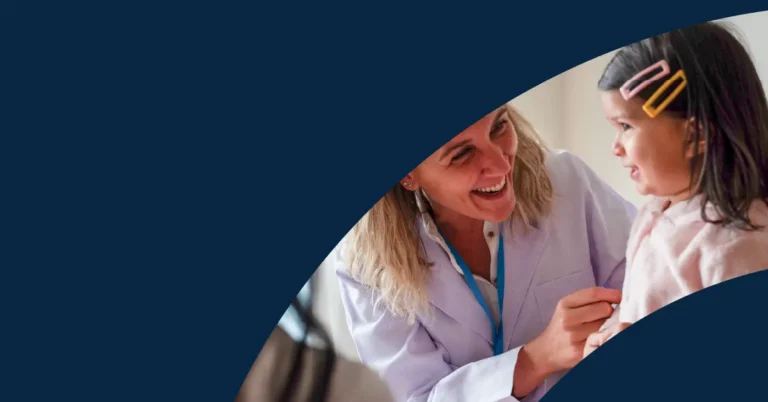 A smiling healthcare worker in a white coat talks to a young girl with hair clips, both interacting warmly; part of the image is covered by a dark blue curved overlay.