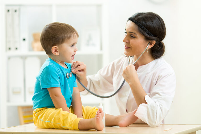 A doctor uses a stethoscope to listen to a smiling young boy's chest during a medical examination in a bright, modern surgery.