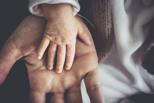 A baby's hand resting on an adult's open palm, highlighting the size difference and conveying a sense of warmth, care, and connection. Both hands are positioned against a soft, white fabric background.