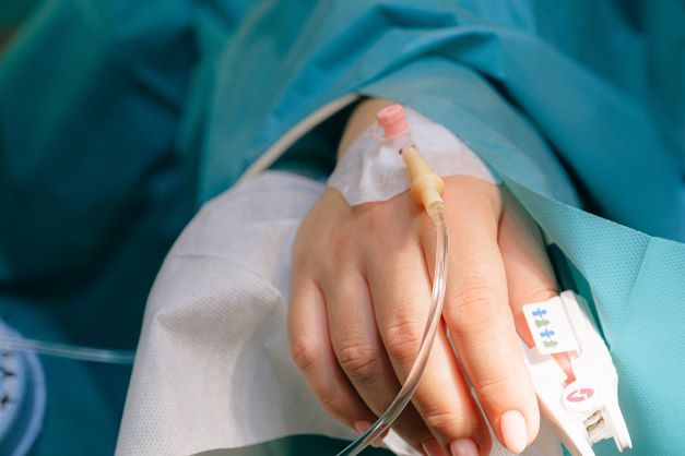 A close-up of a person's hand with an IV cannula inserted, lying on a hospital bed and covered with medical drapes.
