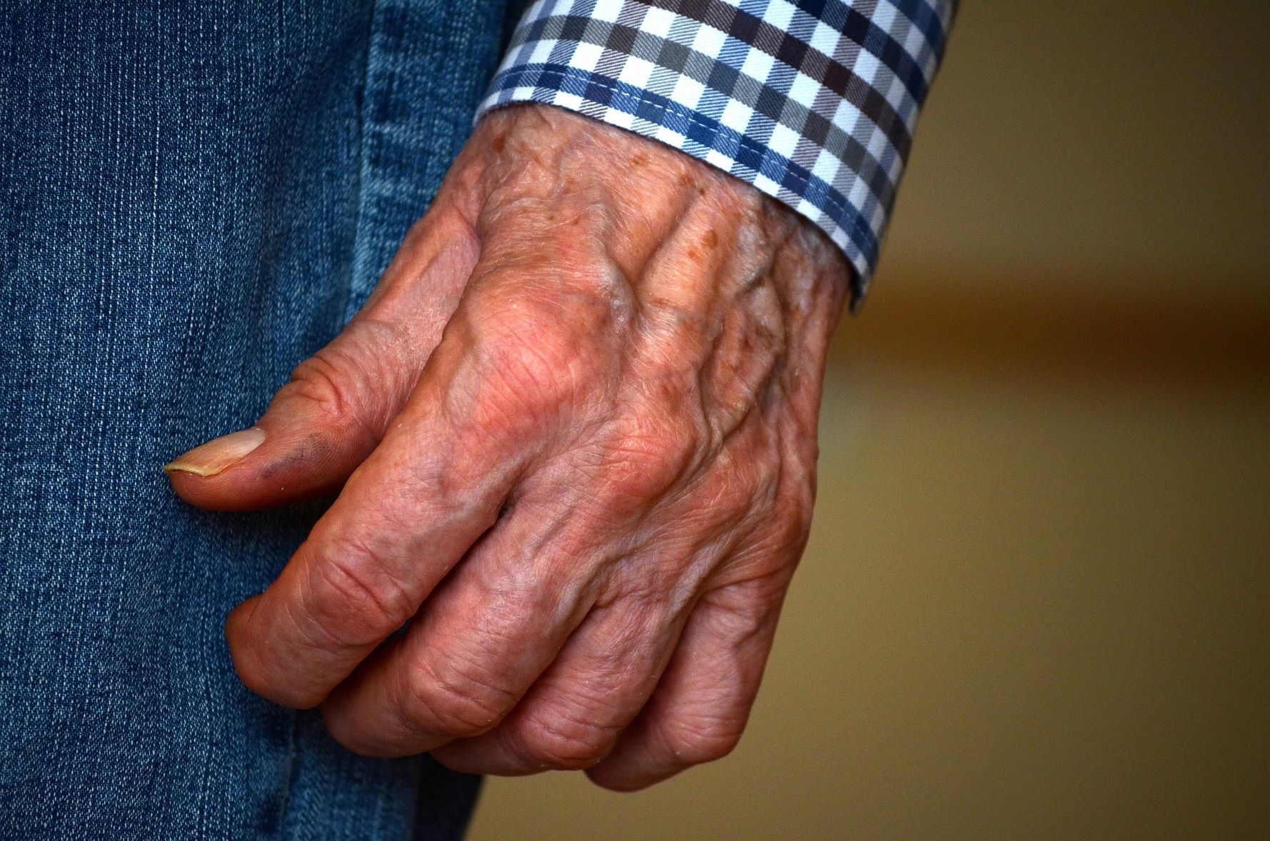 A close-up of an elderly person's hand with visible wrinkles, wearing a blue and white checked shirt and blue jeans, resting by their side.
