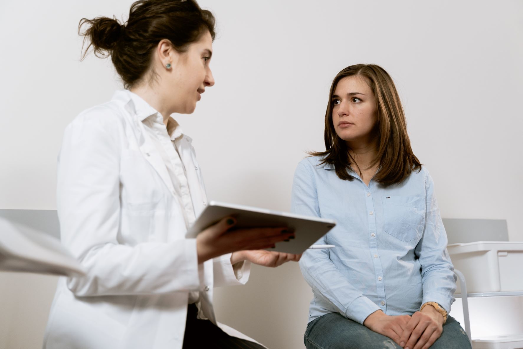 A doctor in a white coat holds a tablet and speaks with a pregnant woman in a light blue blouse, who is sitting and listening attentively in a medical surgery.