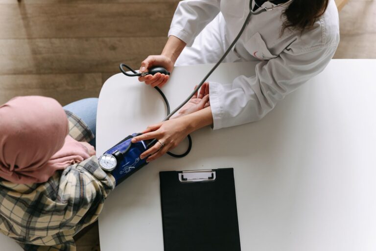 A healthcare professional measures a patient's blood pressure using a cuff and stethoscope at a white table with a clipboard nearby.