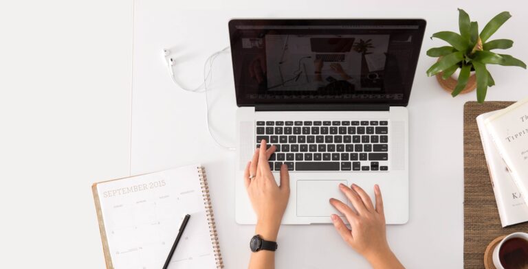 A person types on a laptop at a white desk with a 2015 calendar, pen, plant, and an open book nearby. A charging cable is plugged into the laptop.
