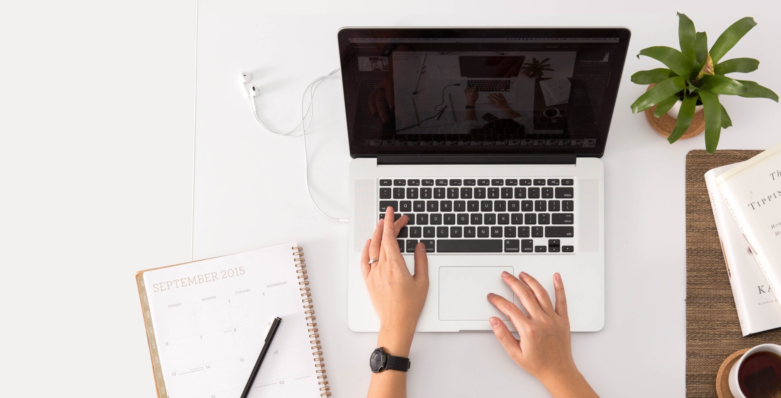 A person types on a laptop at a white desk with a 2015 calendar, pen, plant, and an open book nearby. A charging cable is plugged into the laptop.
