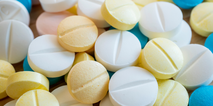 A close-up of assorted round tablets in white, yellow, and blue colours, some with a scored line across the centre, scattered closely together.