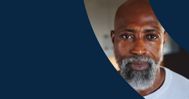 A middle-aged man with a bald head and grey beard looks directly at the camera, with a dark blue graphic partially covering the left side of the image.