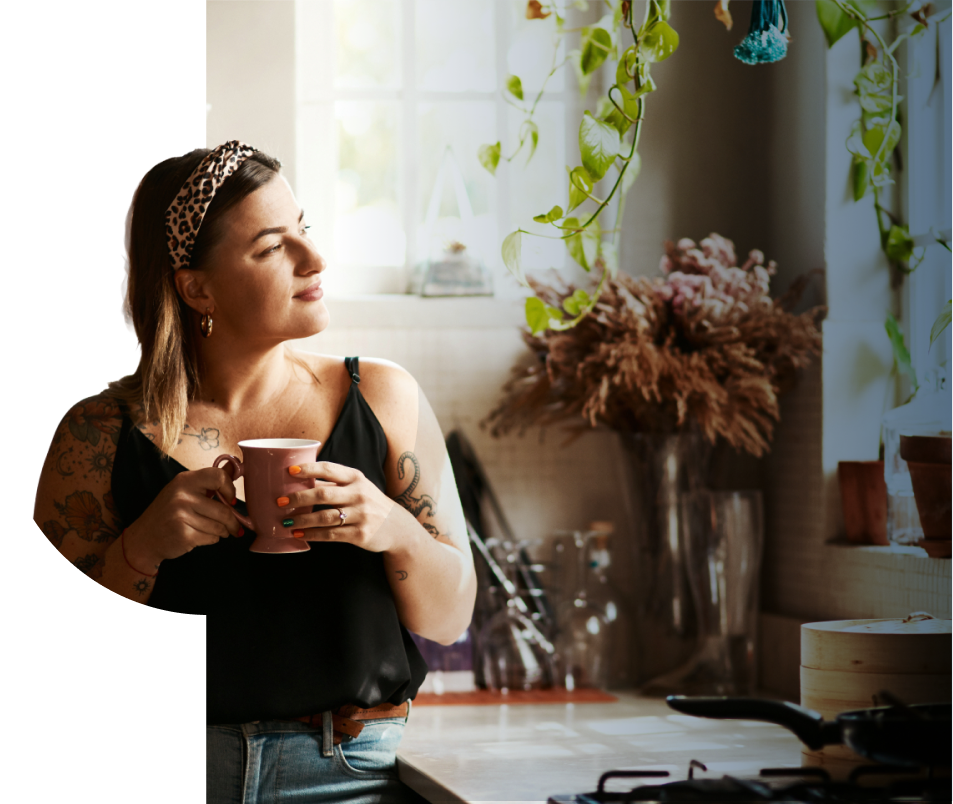 A woman with tattoos holds a mug and looks out of a sunlit kitchen window. She wears a leopard print headband, black top, and jeans. Green plants and dried flowers decorate the cosy kitchen.