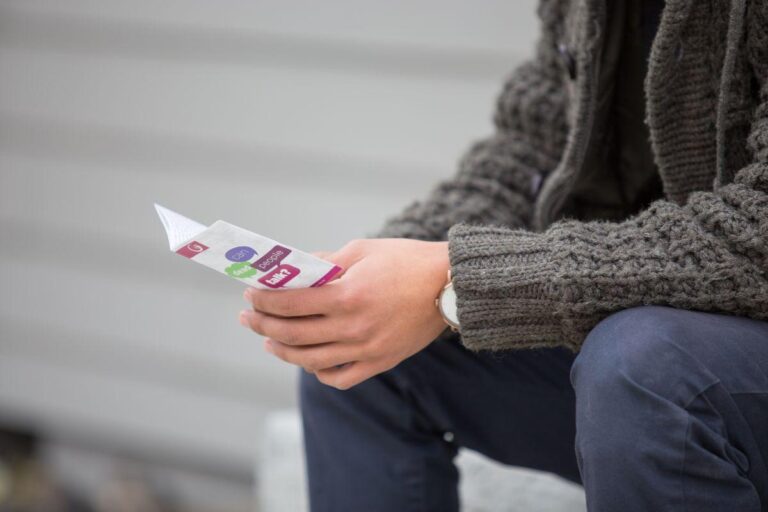 A person in a grey knitted jumper and dark trousers sits outdoors, holding a colourful leaflet or brochure with both hands. The background is blurred, showing horizontal cladding.