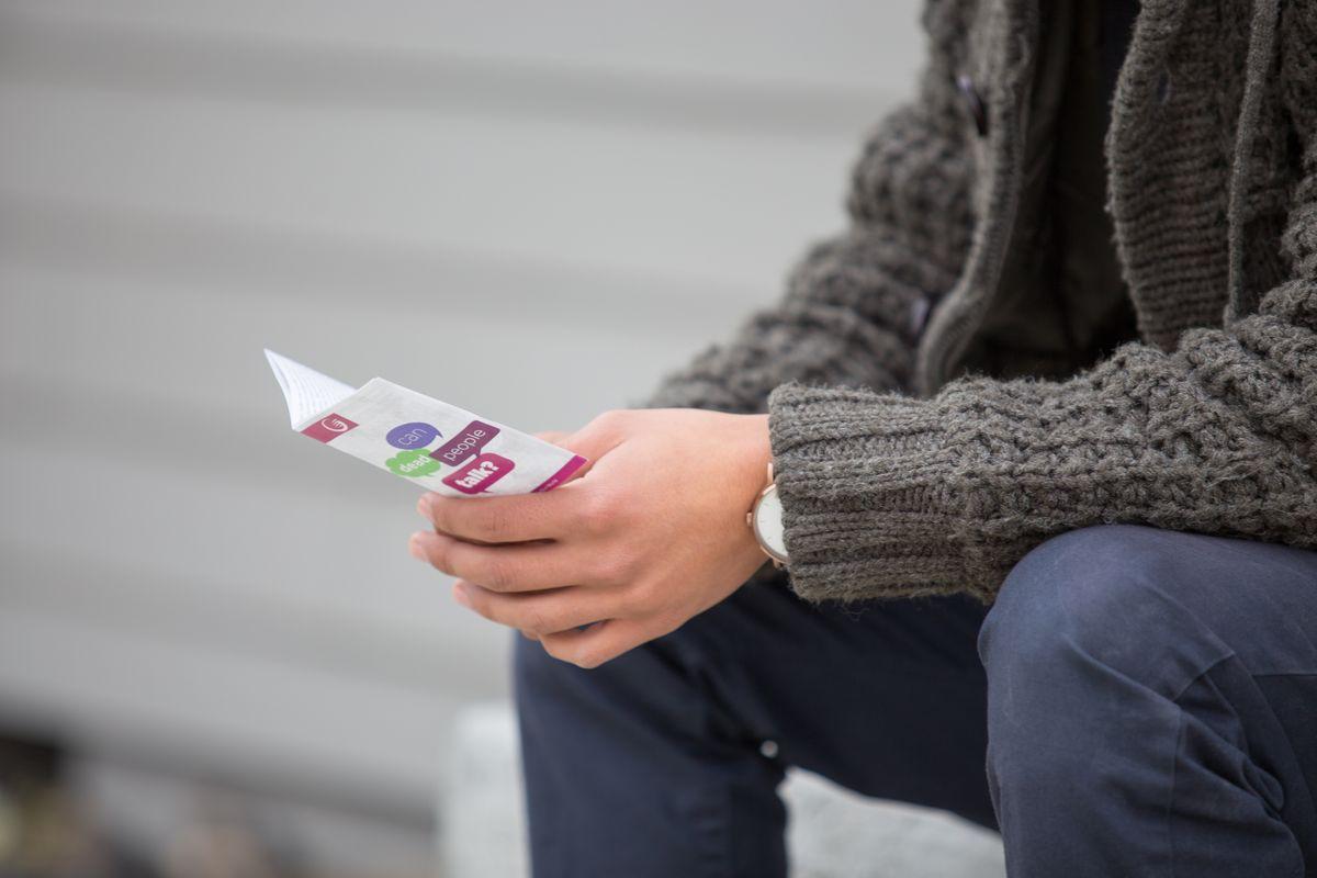 A person in a grey knitted jumper and dark trousers sits outdoors, holding a colourful leaflet or brochure with both hands. The background is blurred, showing horizontal cladding.