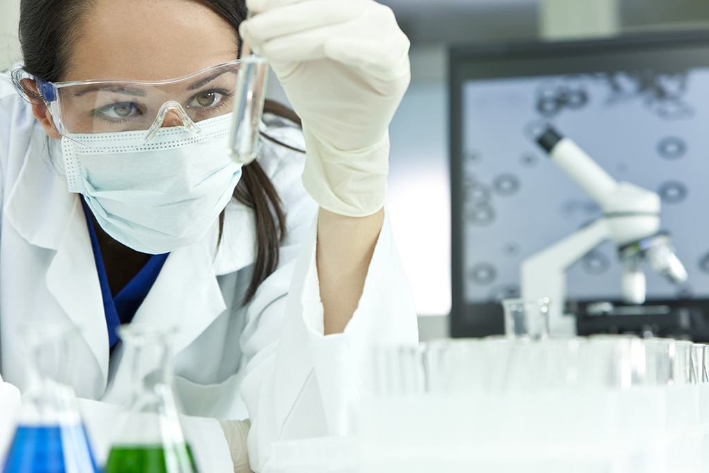 A scientist wearing safety goggles, a mask, and gloves examines a test tube in a laboratory with flasks and a microscope visible in the background.