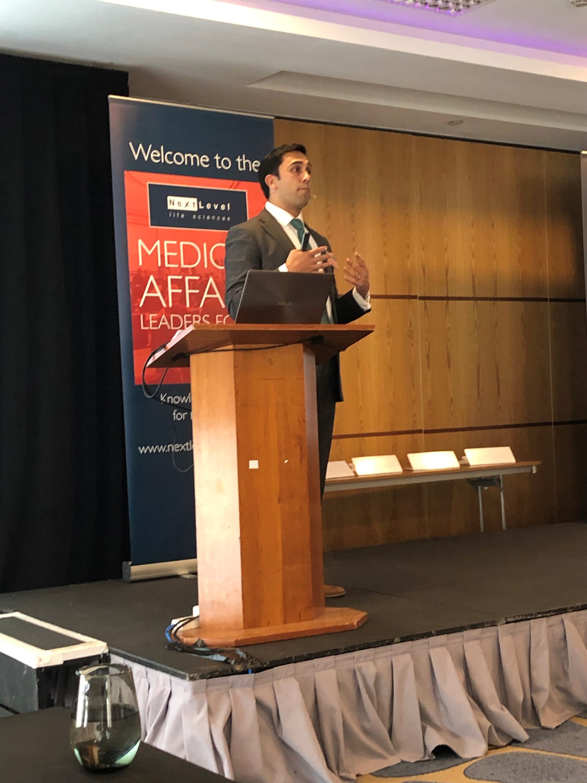 A man in a suit stands at a wooden lectern, speaking at a conference. A banner behind him reads Medical Affairs Leaders Forum. The room has wood panelling and a stage with empty chairs.