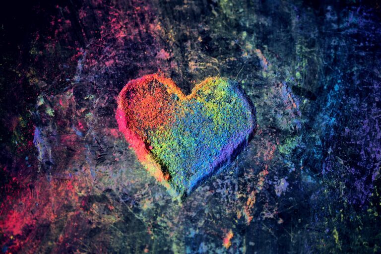 A heart shape made of colourful powdered chalk, featuring a rainbow gradient, on a dark, textured background with scattered chalk dust.