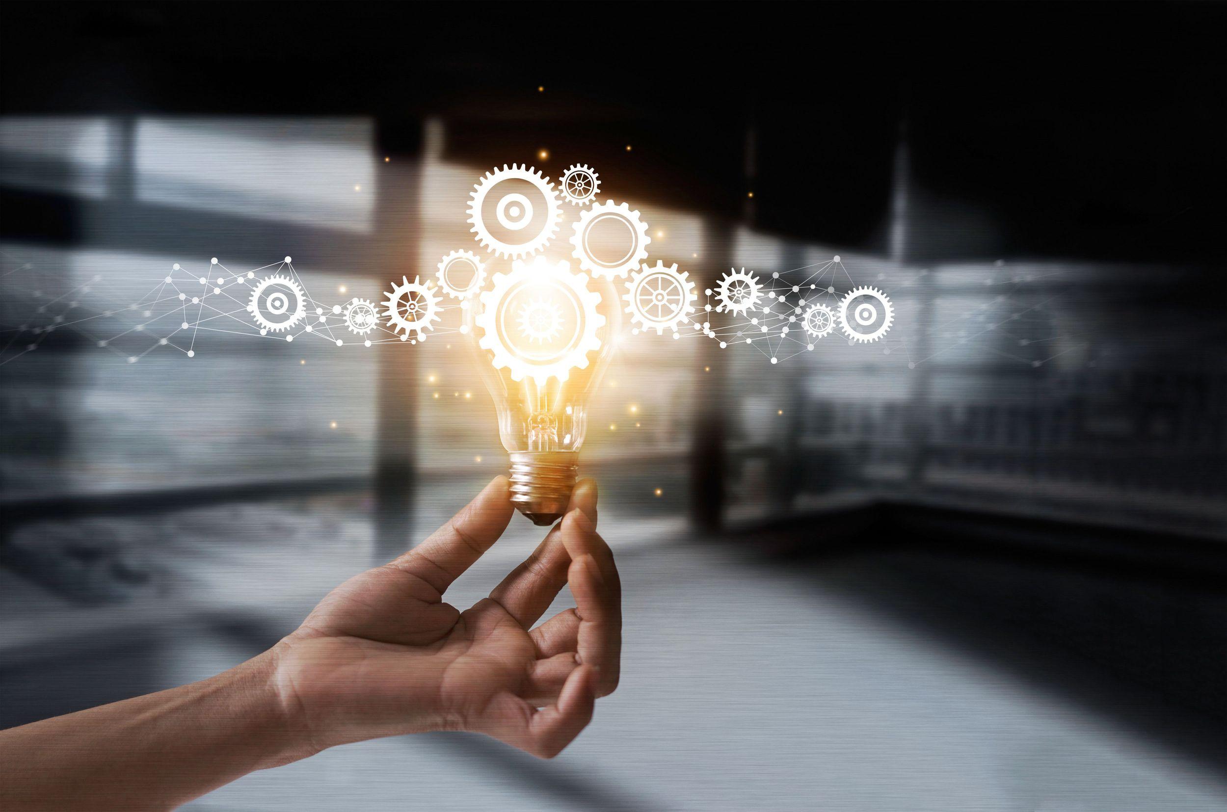 A hand holds a glowing light bulb with gears and network lines above it, symbolising innovation and creative ideas, against a blurred office background.
