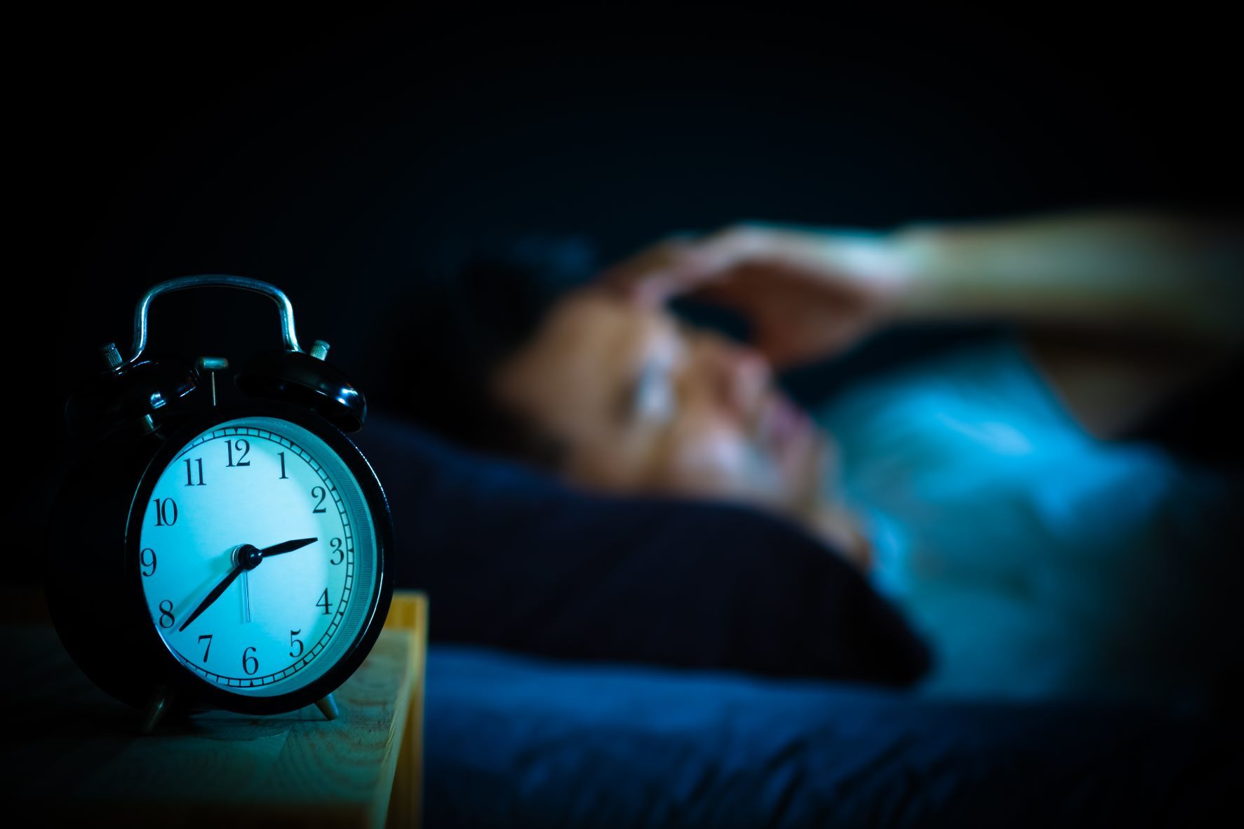 A blurry person lies in bed in the background, appearing stressed. In the foreground, a sharp focus shows an analogue alarm clock on a bedside table, displaying the time as 2:56.