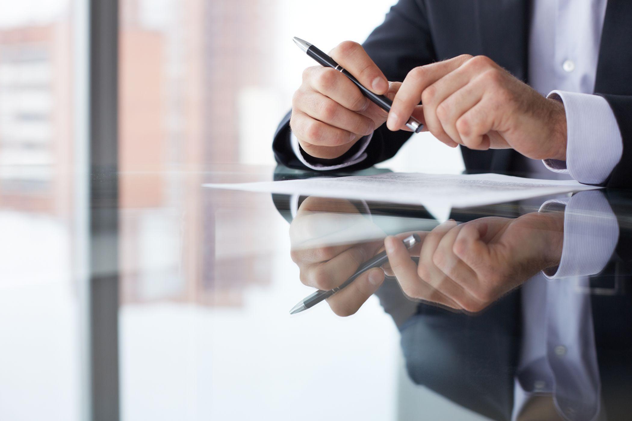 A person in a suit sits at a glass table, holding a pen over a document, with their reflection visible on the table surface.