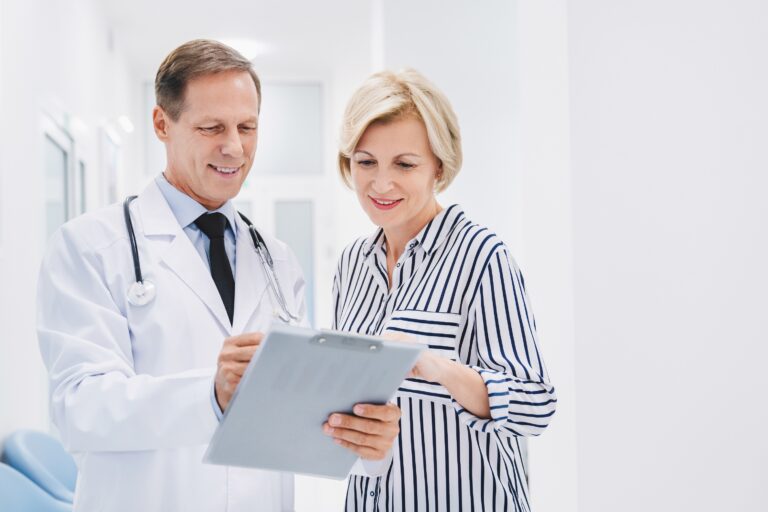 A doctor in a white coat shows a clipboard to a smiling woman in a striped top. They appear to be discussing medical information in a bright, modern clinic setting.