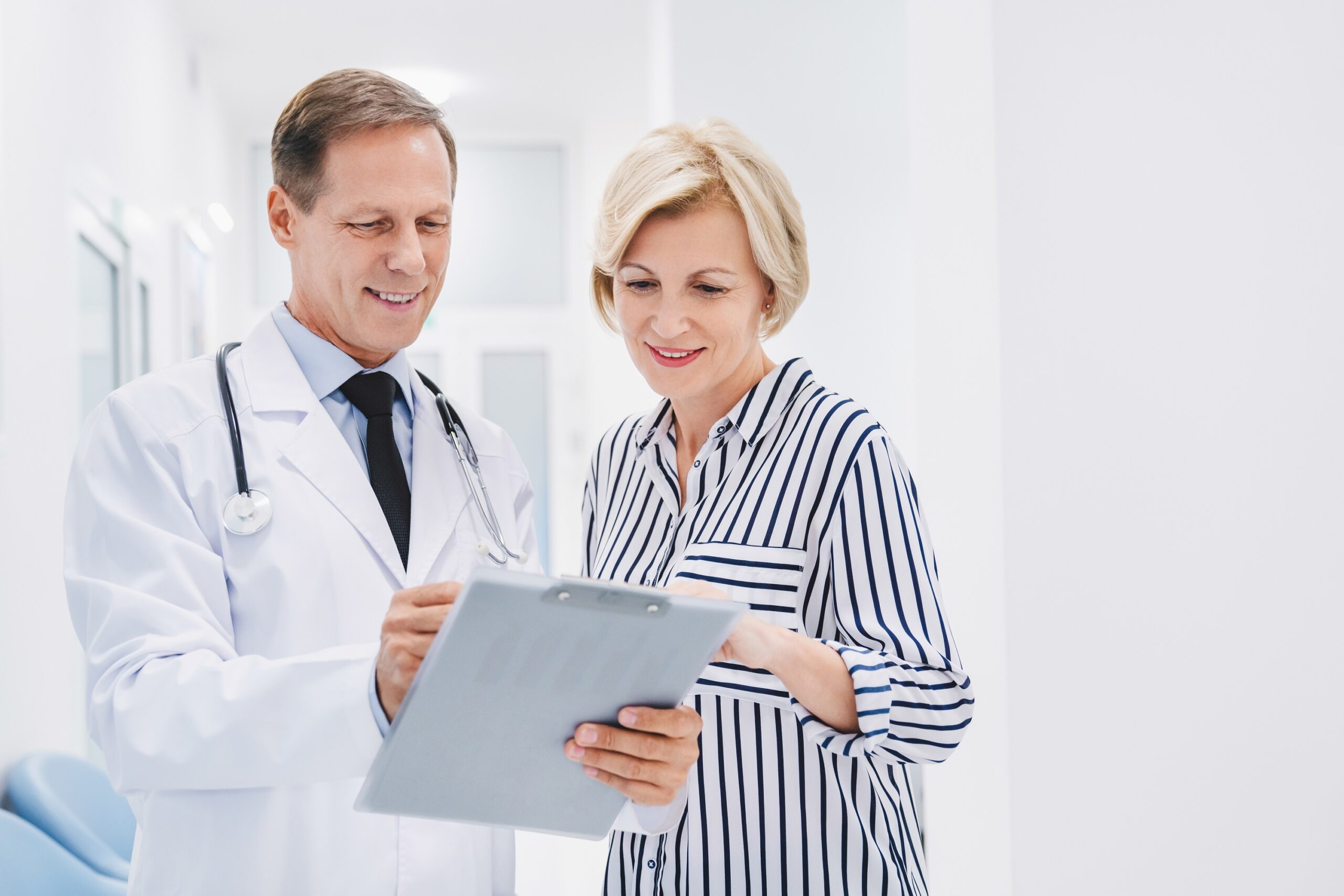 A doctor in a white coat shows a clipboard to a smiling woman in a striped top. They appear to be discussing medical information in a bright, modern clinic setting.