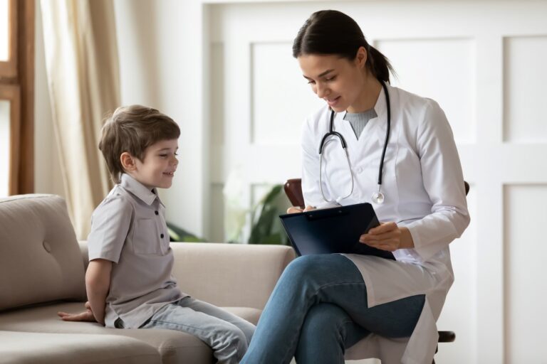 A female doctor with a stethoscope smiles whilst showing a clipboard to a young boy sitting on a sofa in a bright room.