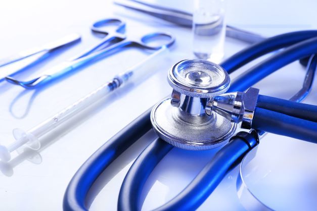 A close-up of medical instruments including a stethoscope, syringe, tweezers, and scissors, all placed on a white surface with a cool blue tint.