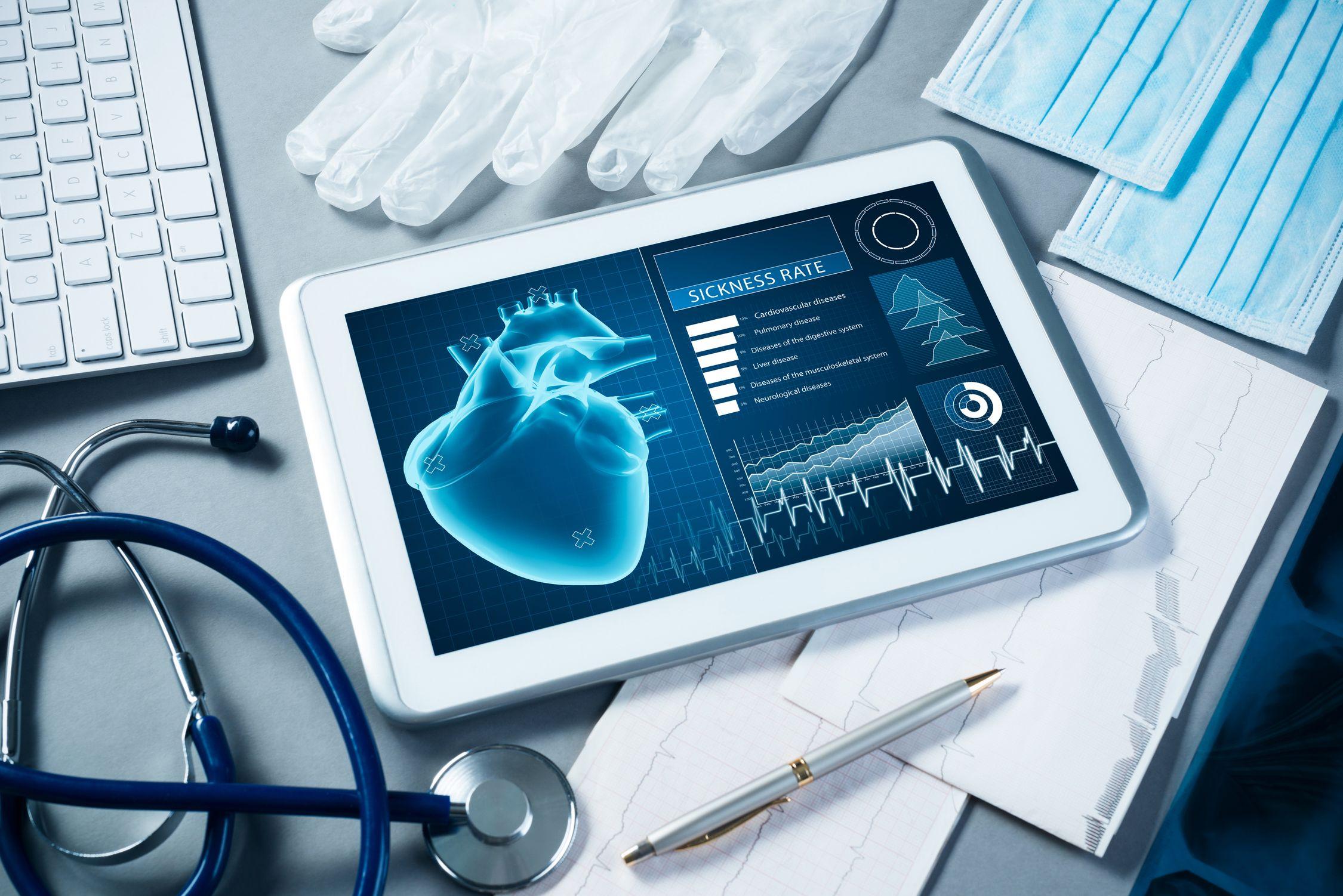 A tablet displays a digital image of a heart and medical stats. Surrounding it are a stethoscope, gloves, face masks, a keyboard, ECG printouts, and a pen, all on a grey surface.