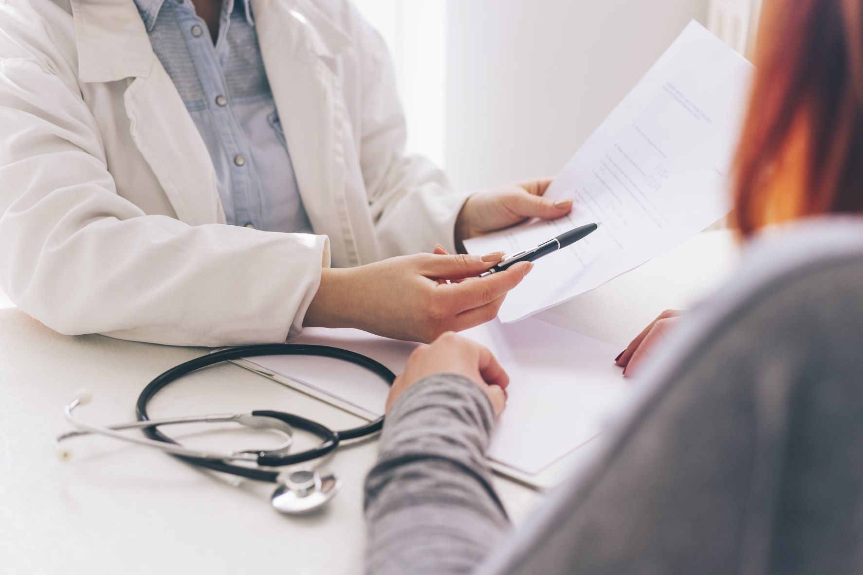 A doctor in a white coat explains a document to a patient. The doctor points to the paper with a pen, and a stethoscope lies on the desk. Only their hands and torsos are visible.
