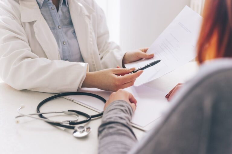 A doctor points to a form with a pen whilst discussing it with a patient. A stethoscope lies on the desk in front of them.