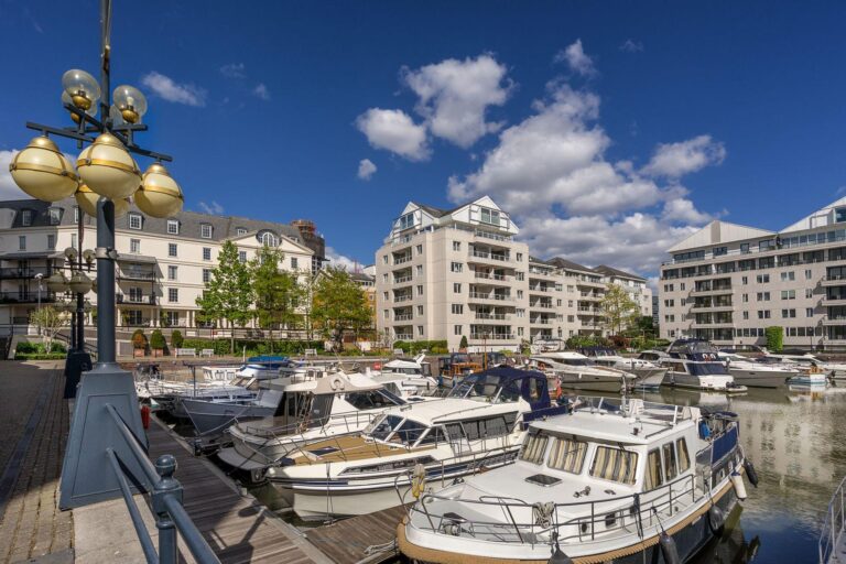 Boats are moored at a marina surrounded by modern blocks of flats under a blue sky with scattered clouds. A lamp-post is visible in the foreground on the left.