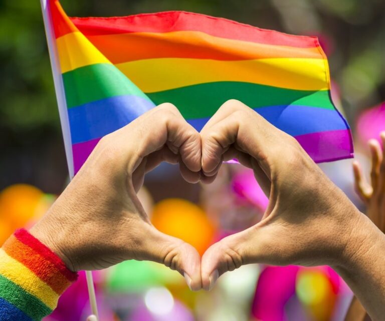 Two hands form a heart shape in front of a waving rainbow pride flag. The person wears a rainbow wristband, symbolising LGBTQ+ pride and support at what appears to be a colourful outdoor event.
