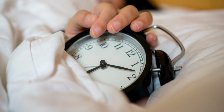 A hand rests on a black alarm clock partially covered by a white duvet, suggesting someone has just woken up or is about to silence the alarm.